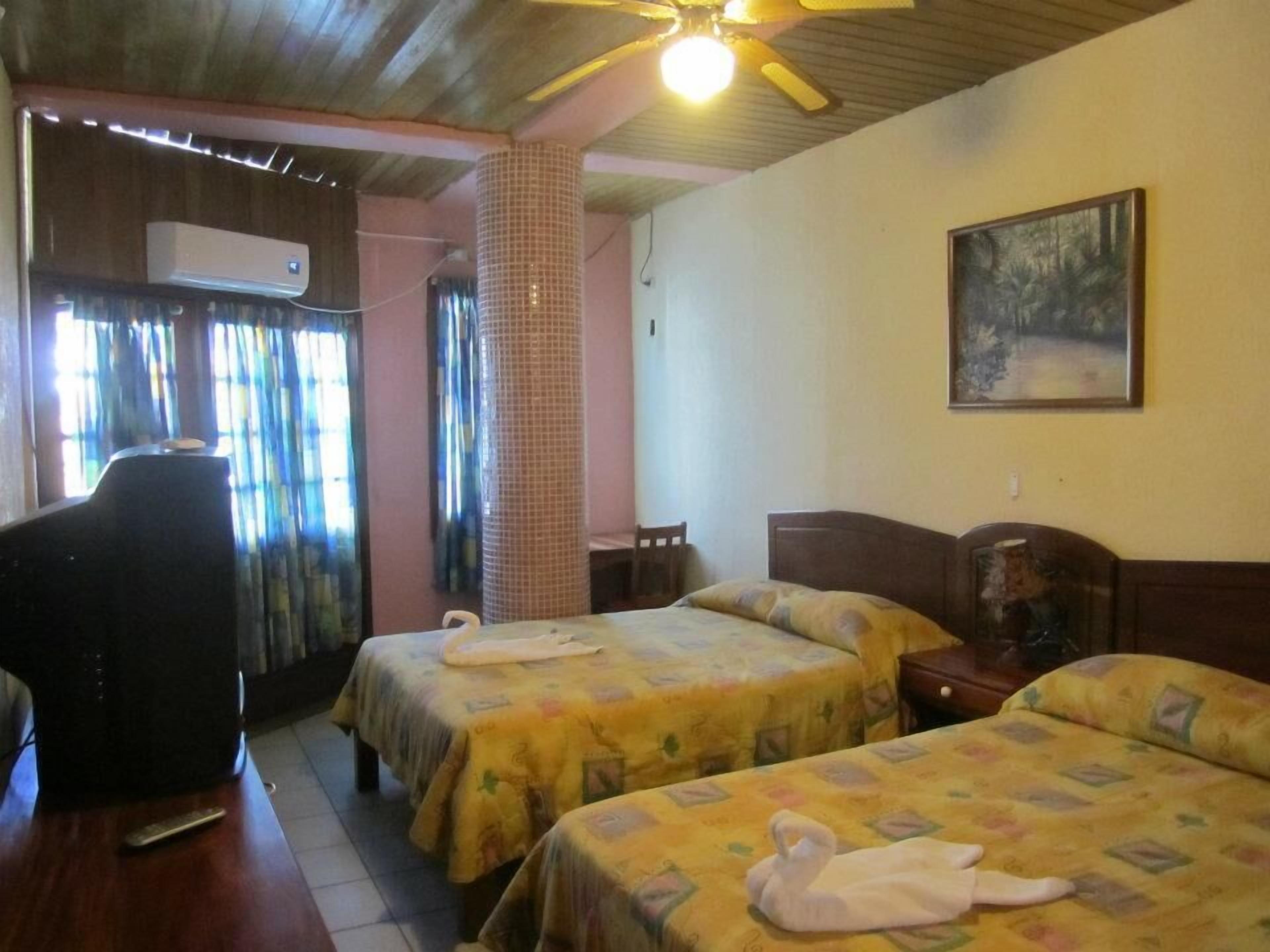 A hotel room with two beds, a desk, a chair, an air conditioning unit, and a ceiling fan.