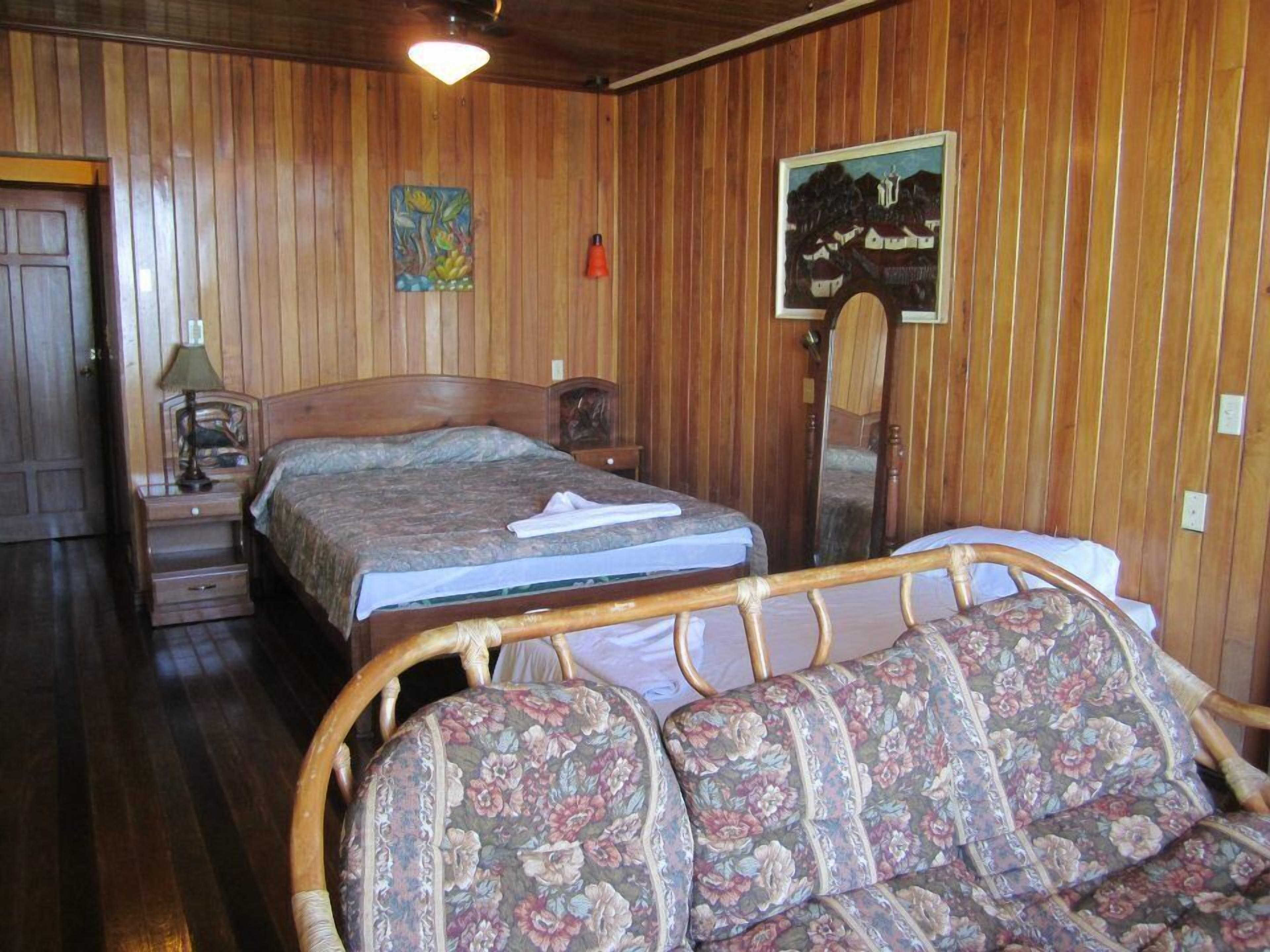 A wooden-paneled room with a bed, a chair, a nightstand, a lamp, and two paintings on the wall.