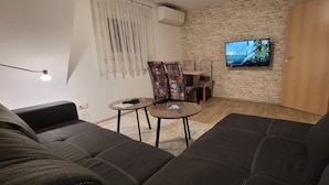 Un salon avec un mur en pierre, un téléviseur fixé au mur, un canapé, une petite table avec une télécommande et une lampe.