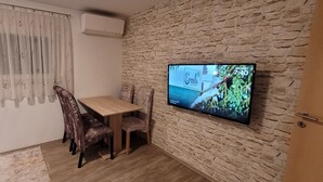Un coin repas avec une table et des chaises en bois, un téléviseur fixé à un mur en pierre et une fenêtre ornée de rideaux légers.