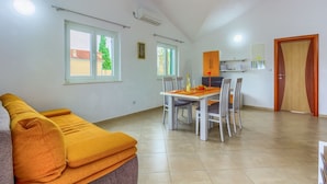 A spacious living area with a sofa, dining table, and chairs. There are windows allowing natural light, an air conditioning unit, and a door with a unique design.