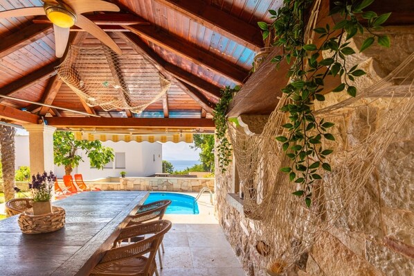 A covered outdoor area with a pool, wooden furniture, and stone walls.