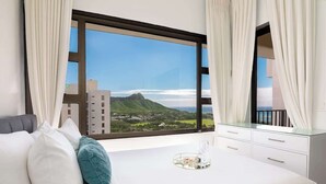A hotel room with a large window, a bed, a bedside table with a tray, and a view of a mountain and buildings. - Ocean-view condo w/ pool near Waikiki Beach (Honolulu)