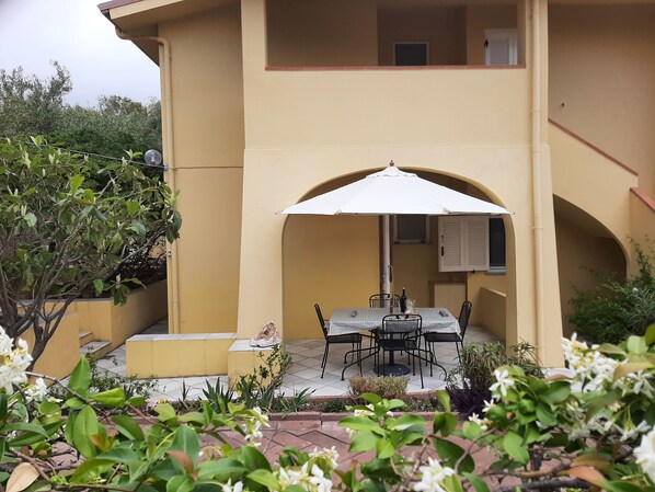 An outdoor patio with a table and chairs under a white umbrella, surrounded by greenery and a yellow building.