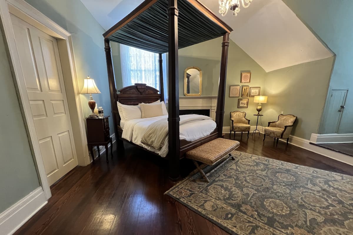 Un lit à baldaquin en bois, une petite table de chevet avec une lampe, une chaise et un tapis à motifs dans une chambre dotée d’un plancher en bois et d’un plafond en pente.