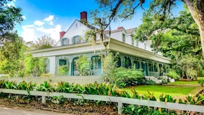 A large, two-story house with a white facade, green shutters, and a porch with ornate railings.