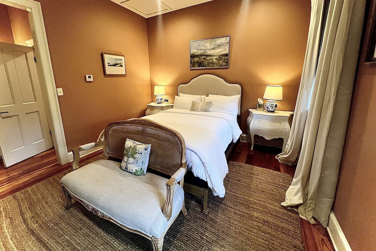 Une chambre d’hôtel avec un grand lit, deux tables de chevet avec des lampes, un fauteuil avec un coussin, un tableau au mur et une porte.
