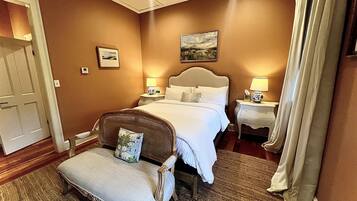 Una habitación de hotel con una cama grande, dos mesitas de noche con lámparas, una silla con cojín, un cuadro en la pared y una puerta.