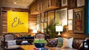 A cozy room with a wooden wall, a yellow sign with the name 'Elta', a brown leather sofa, a wooden coffee table, and a potted plant.