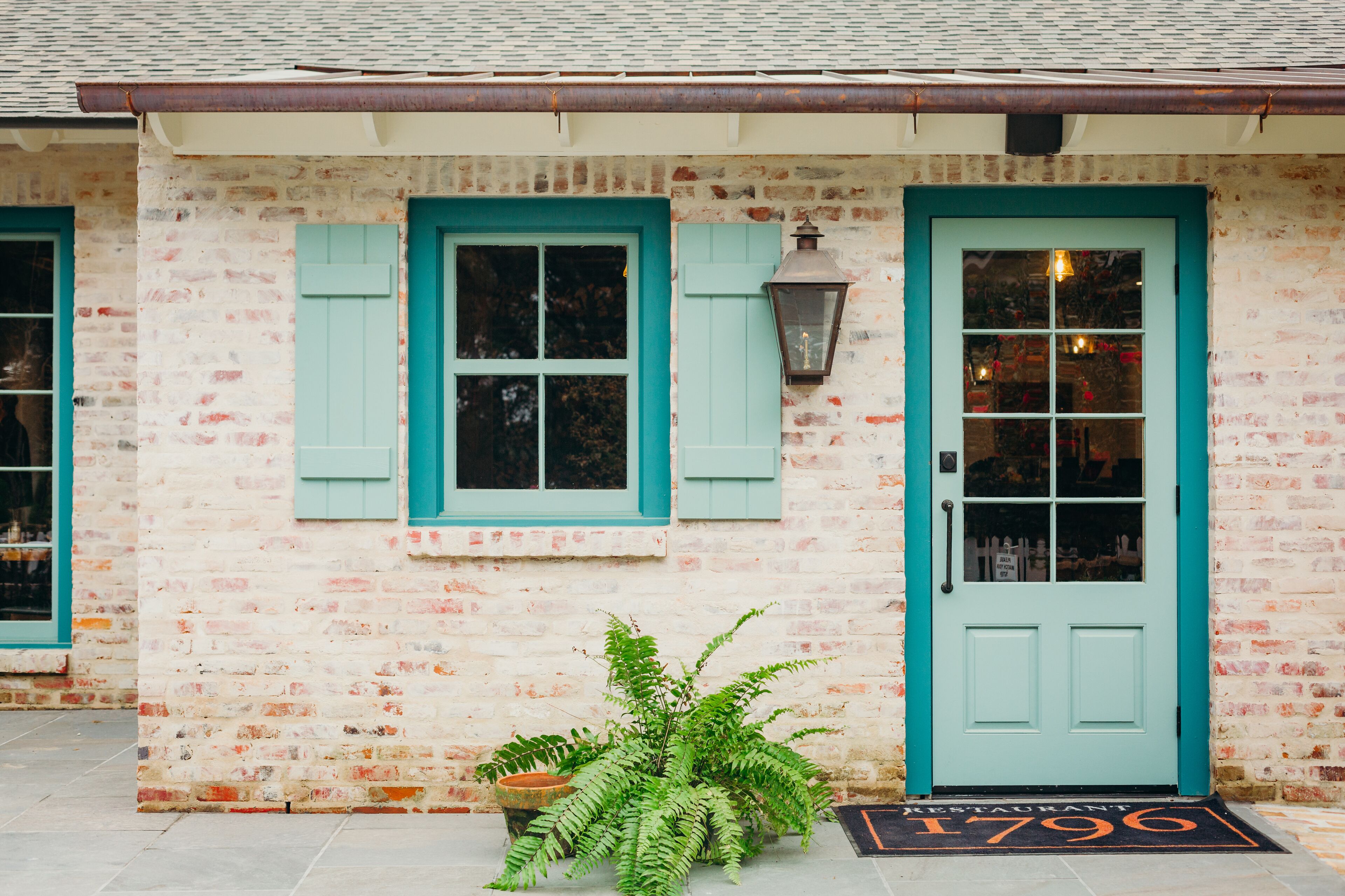 A building with a white brick facade, teal shutters, and a door with a glass panel.