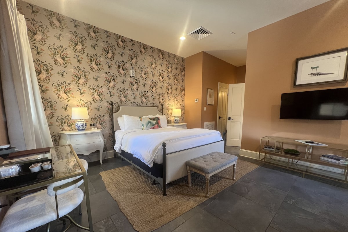 Une chambre d’hôtel avec un grand lit, deux tables de chevet avec des lampes, un banc, un bureau avec une chaise, un téléviseur à écran plat et une œuvre d’art accrochée au mur.