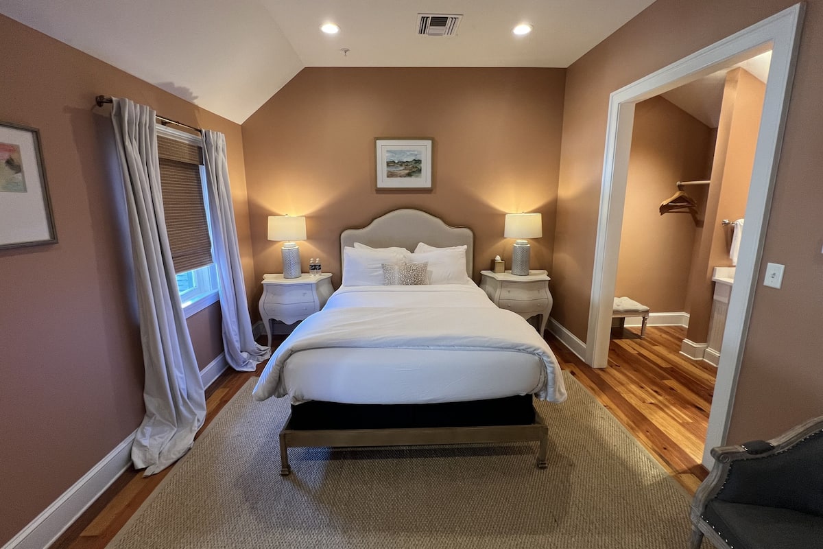 Une chambre à coucher bien rangée, avec un grand lit, deux tables de chevet avec des lampes, un tableau encadré au-dessus du lit et une porte donnant sur une autre pièce.
