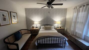 Ein Schlafzimmer mit einem Bett, zwei Nachttischen mit Lampen, einem Deckenventilator, einem Fenster mit Vorhängen und zwei gerahmten Bildern an der Wand.