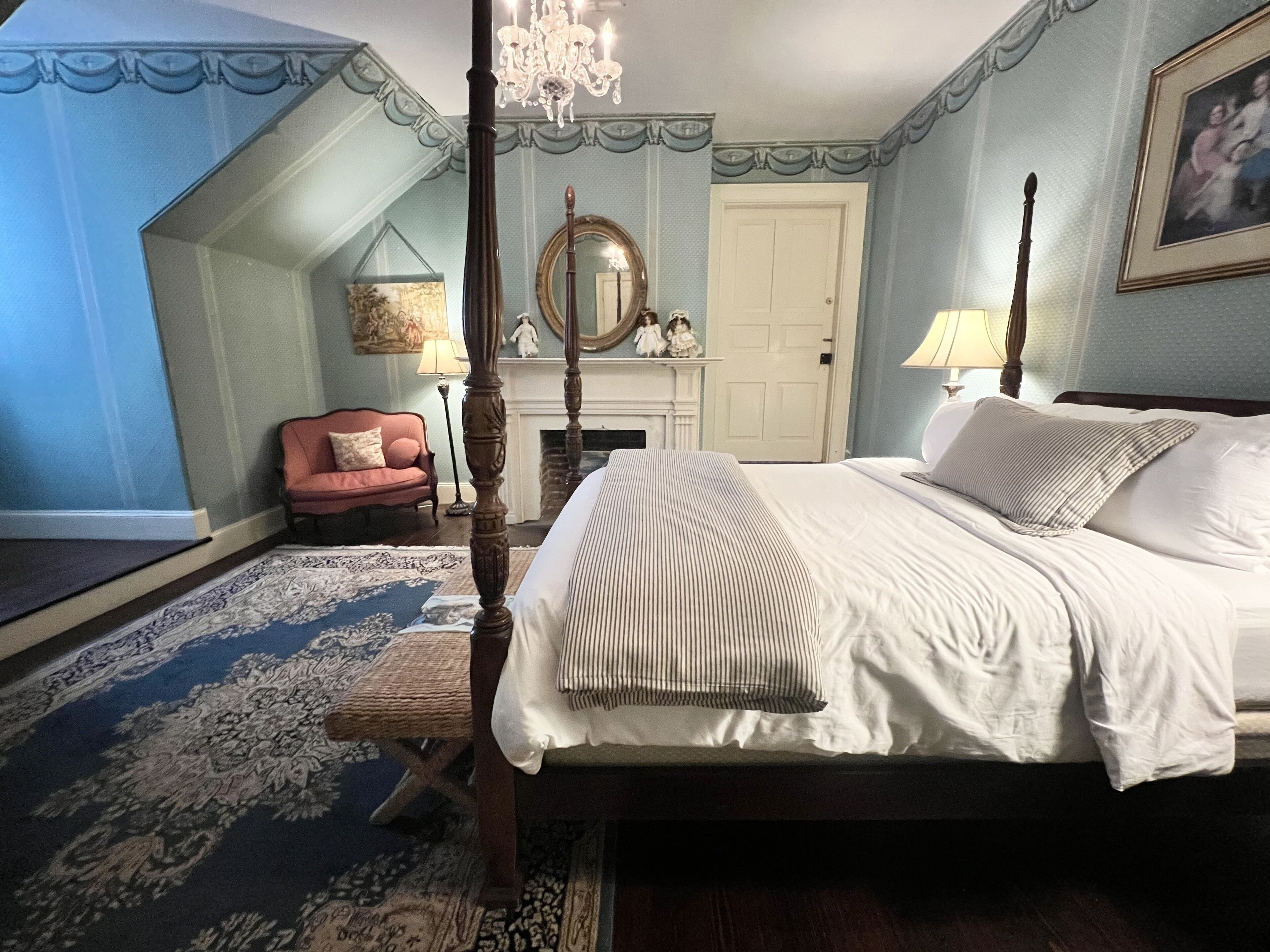 A bedroom with a four-poster bed, a chandelier, a fireplace, and a large mirror.