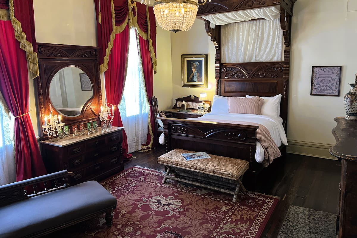 Une chambre à coucher de style traditionnel, avec un lit à baldaquin, un lustre et une fenêtre ornée de rideaux rouges et dorés.