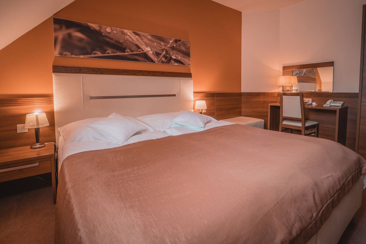 Habitación de hotel con una cama grande, dos mesitas de noche con lámparas, un cabecero de madera, un escritorio pequeño con una silla y un espejo.