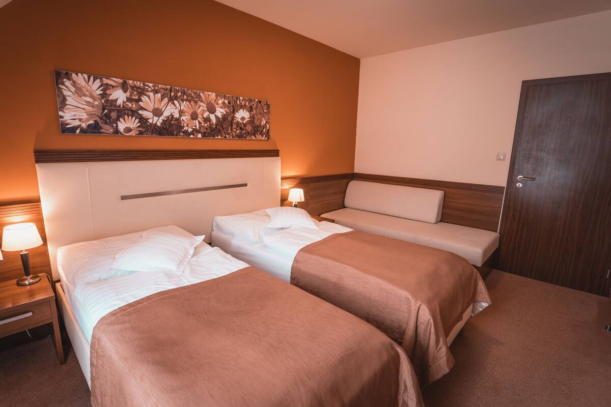 Habitación de hotel con dos camas, cabecera de madera, mesita de noche con lámpara, una puerta y una obra de arte en la pared.