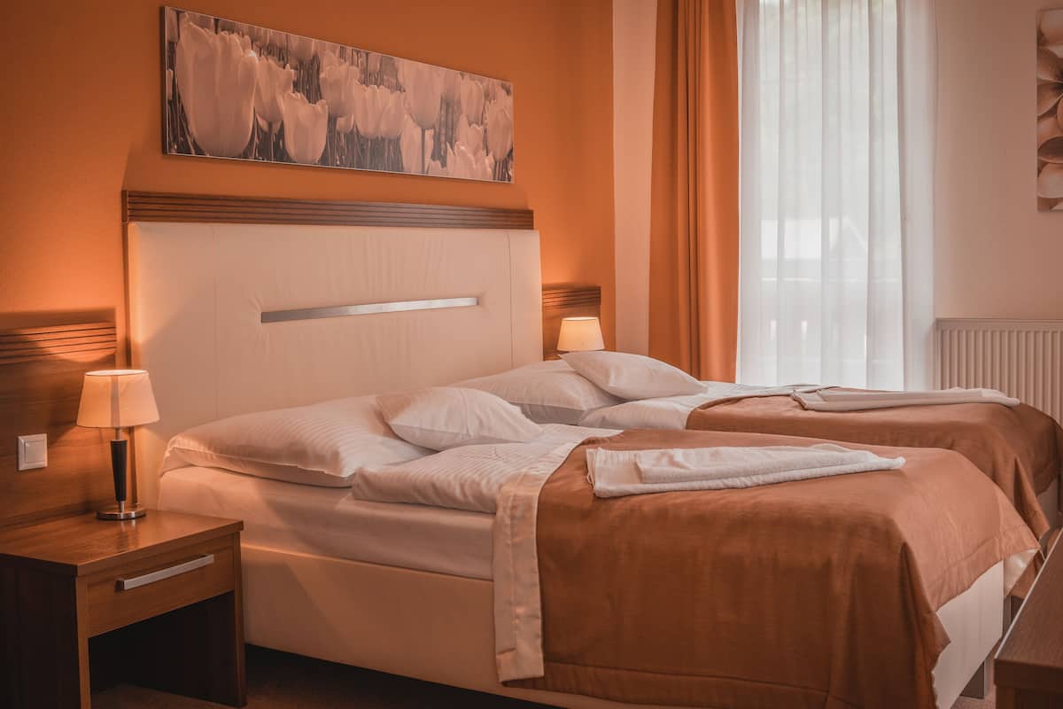Una habitación de hotel con dos camas, cabecero de madera, mesita de noche con lámpara, radiador y un cuadro en la pared.
