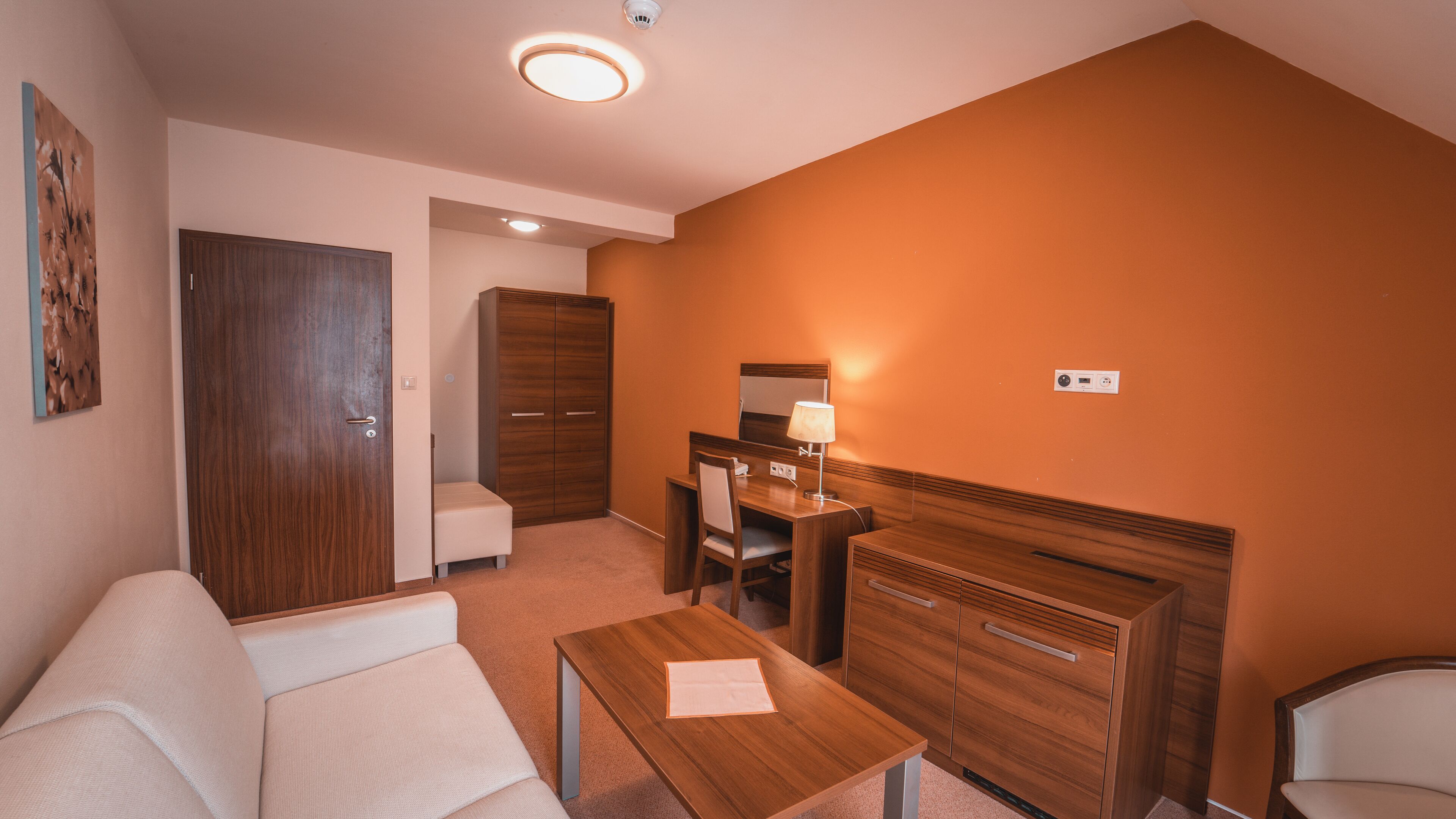 Une chambre d’hôtel avec un canapé, un bureau en bois avec une lampe, une chaise, une petite table, une armoire et un miroir.