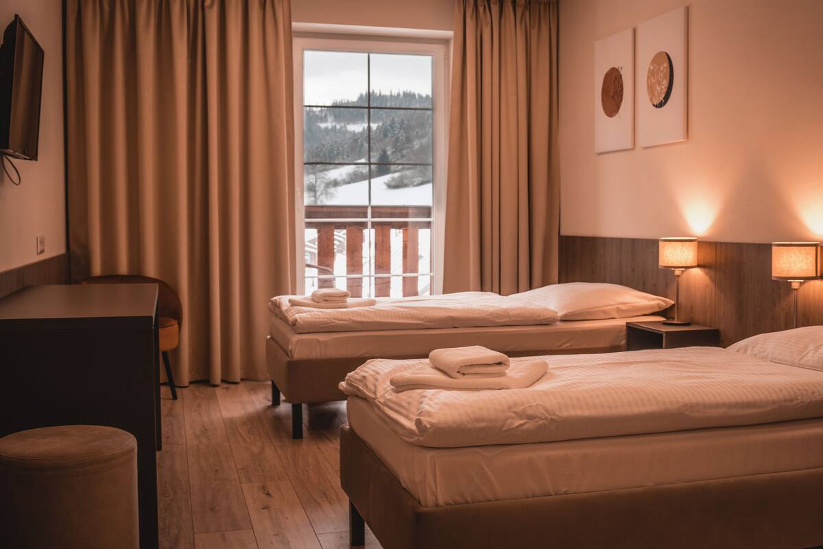 Habitación de hotel con dos camas, un escritorio, una silla, una lámpara y una ventana con vista a un paisaje nevado.