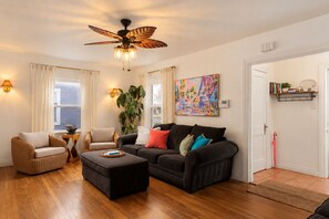 Un salon avec un ventilateur de plafond, un canapé noir, un fauteuil marron, une table basse en bois et un tableau accroché au mur.