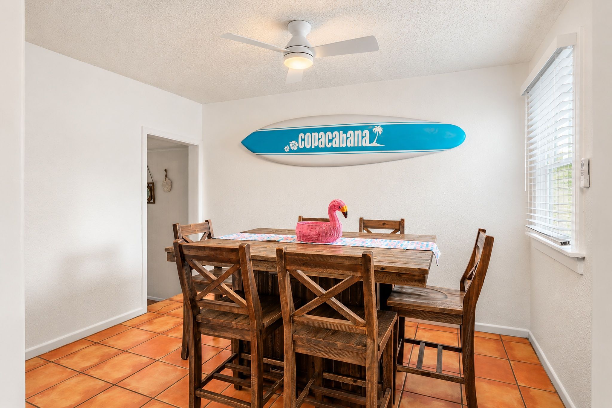 Un coin repas avec une table et des chaises en bois, un ventilateur de plafond, un panneau décoratif mural et une fenêtre avec des stores.