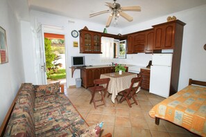 A compact kitchen with wooden cabinets, a dining table with chairs, a sofa, and a ceiling fan.