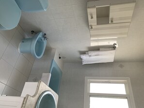 Casa de banho com sanita, bidé e banheira. Há um porta-toalhas com uma toalha branca e uma janela com persianas.