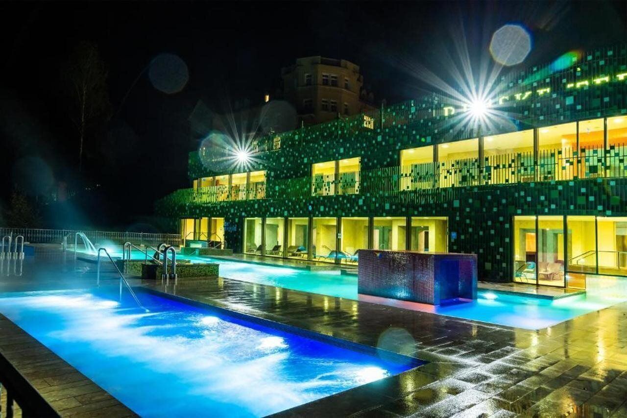 Une piscine agrémentée d’un élément décoratif aquatique, un bâtiment au design moderne et une ambiance nocturne.