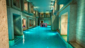 Una piscina interior larga y estrecha, con una chimenea central, rodeada de paredes de piedra y múltiples ventanas.