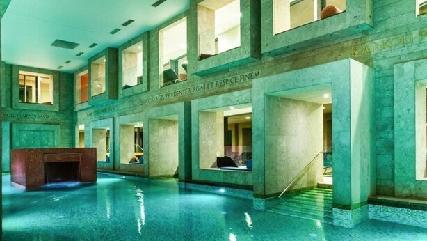 Una amplia piscina interior con agua cristalina, rodeada de muros de piedra y varias ventanas.