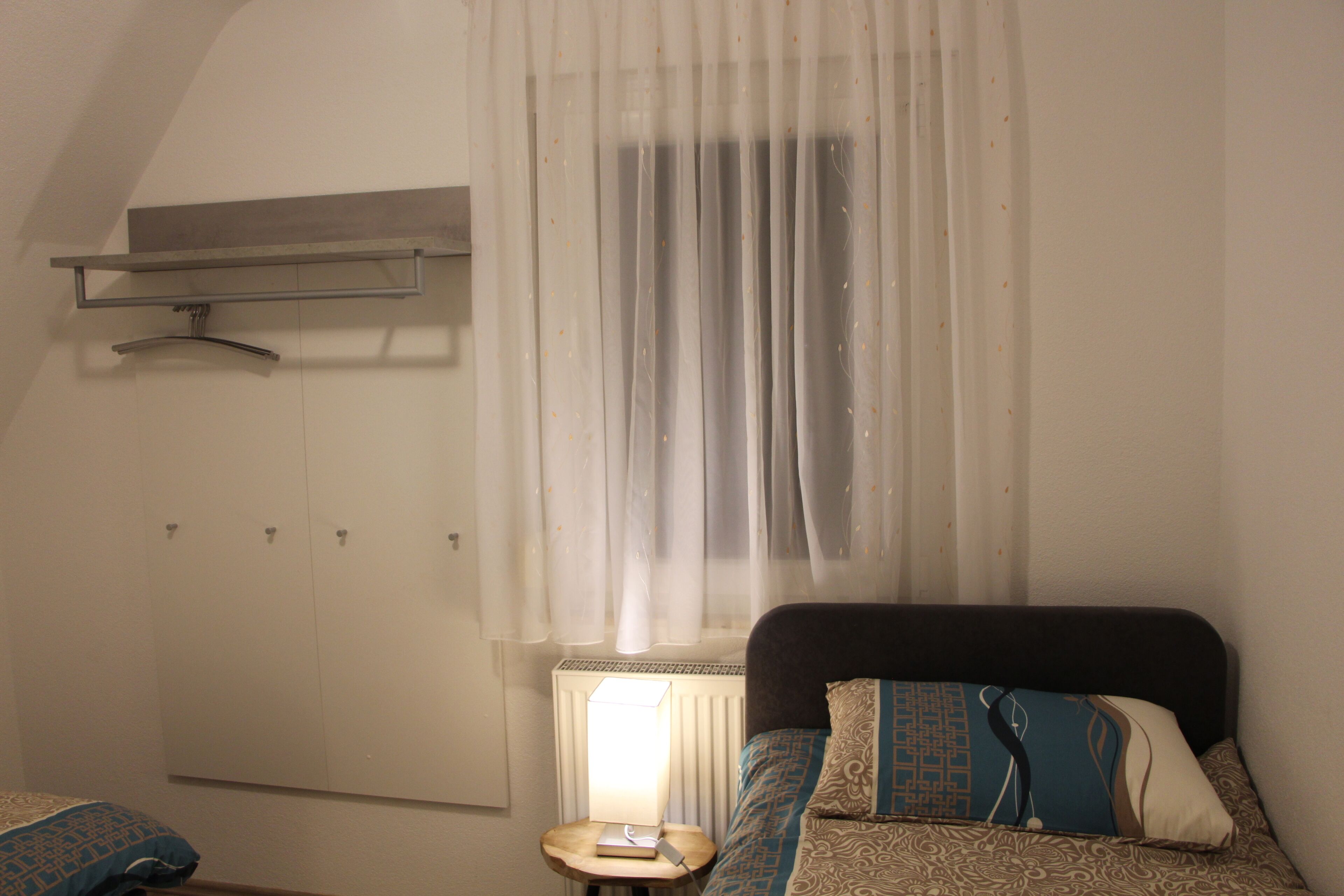 A bedroom with a bed, a bedside table with a lamp, a radiator, and a window with sheer curtains.