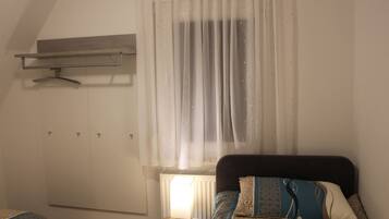 A bedroom with a bed, a bedside table with a lamp, a radiator, and a window with sheer curtains.