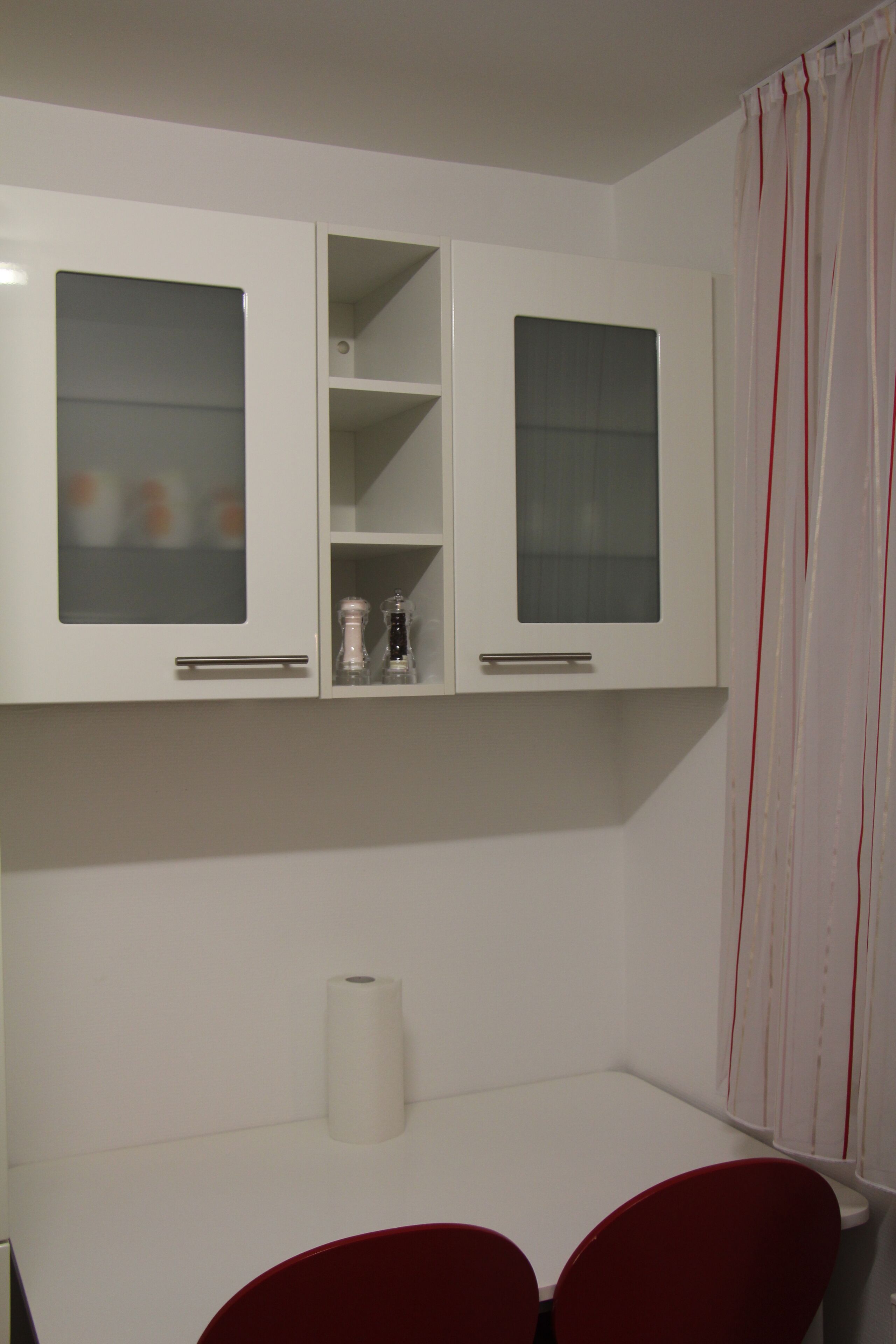 Une pièce avec des meubles blancs aux portes vitrées, une chaise rouge et une table blanche.