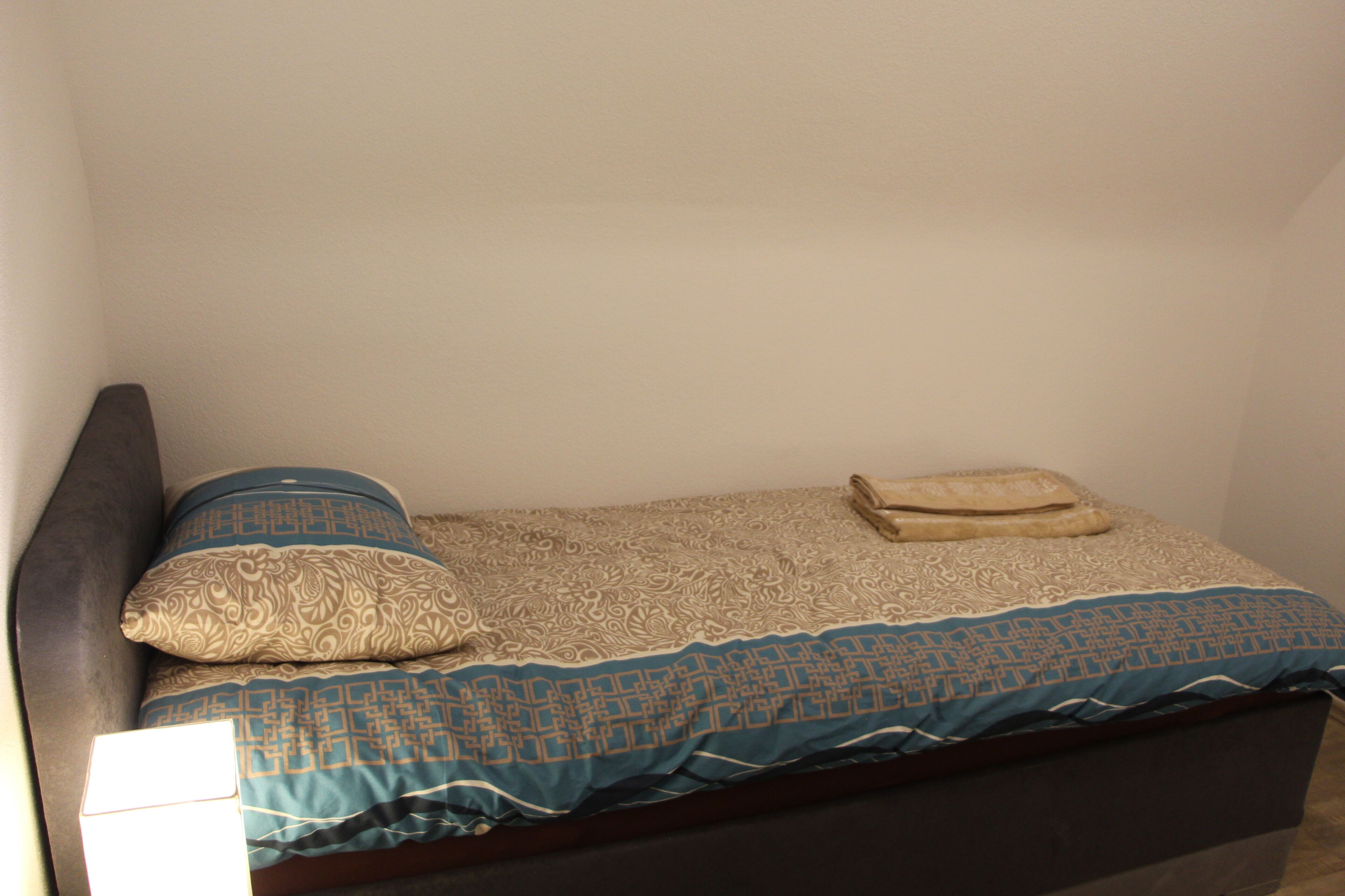 Une petite chambre avec un lit simple, dotée d’une tête de lit sombre, d  ’une couvre-lit à motifs et de deux oreillers.