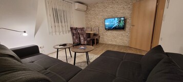 Uma sala de estar com parede de pedra, uma televisão fixada nela, um sofá, uma mesinha de centro com um controle remoto e um abajur.