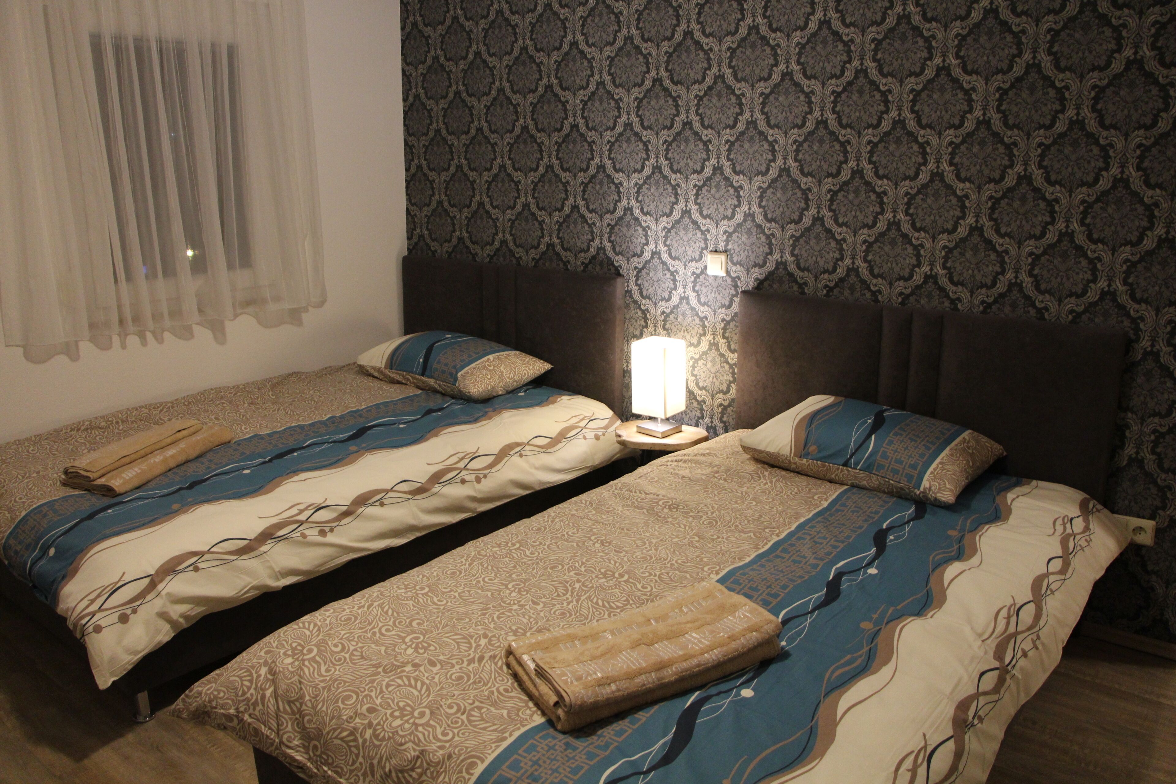 Ein Hotelzimmer mit zwei Betten, jeweils mit gemusterten Bettbezügen und Kissen, einem Nachttisch mit Lampe sowie tapizierten Wänden.