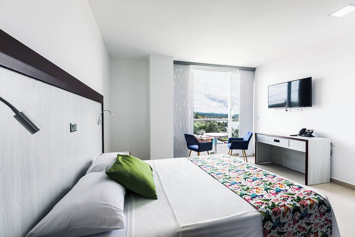 A modern hotel room with a bed, a desk, a chair, a TV, and a balcony with a view.