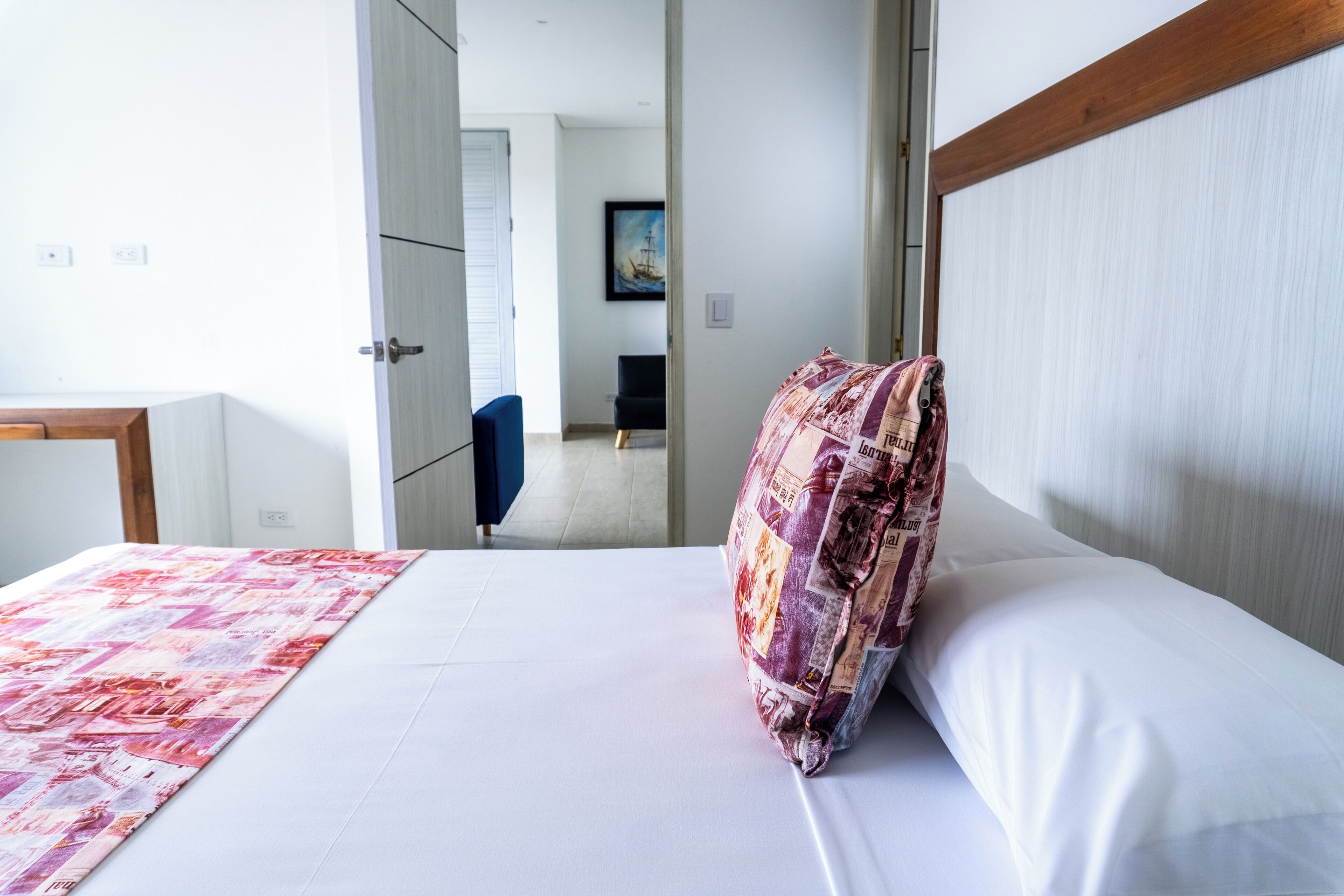 A hotel room with a bed, a pillow, and a blanket. There is a door leading to another room and a hallway with a chair.