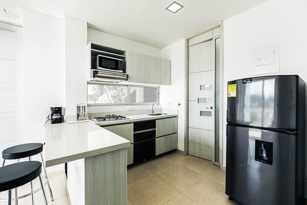 A modern kitchen with a stainless steel refrigerator, a microwave, a gas stove, and a sink. There are two black bar stools and a window with blinds. - Hotel Santorini (Guatapé)