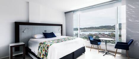 Una habitación de hotel moderna con una cama grande, una mesita, dos sillas y una vista panorámica de la ciudad a través de amplios ventanales.