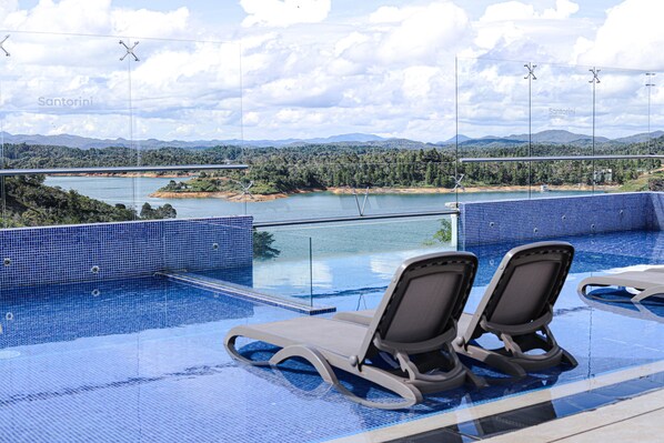 Uma piscina infinita com duas espreguiçadeiras e vista para um lago e montanhas.