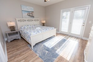 A bedroom with a bed, two bedside tables, a nightstand, a painting, and a door with blinds. - FADE AWAY -New 5 Bedroom House Only Steps From The Ocean With a Private Pool (Surfside Beach)