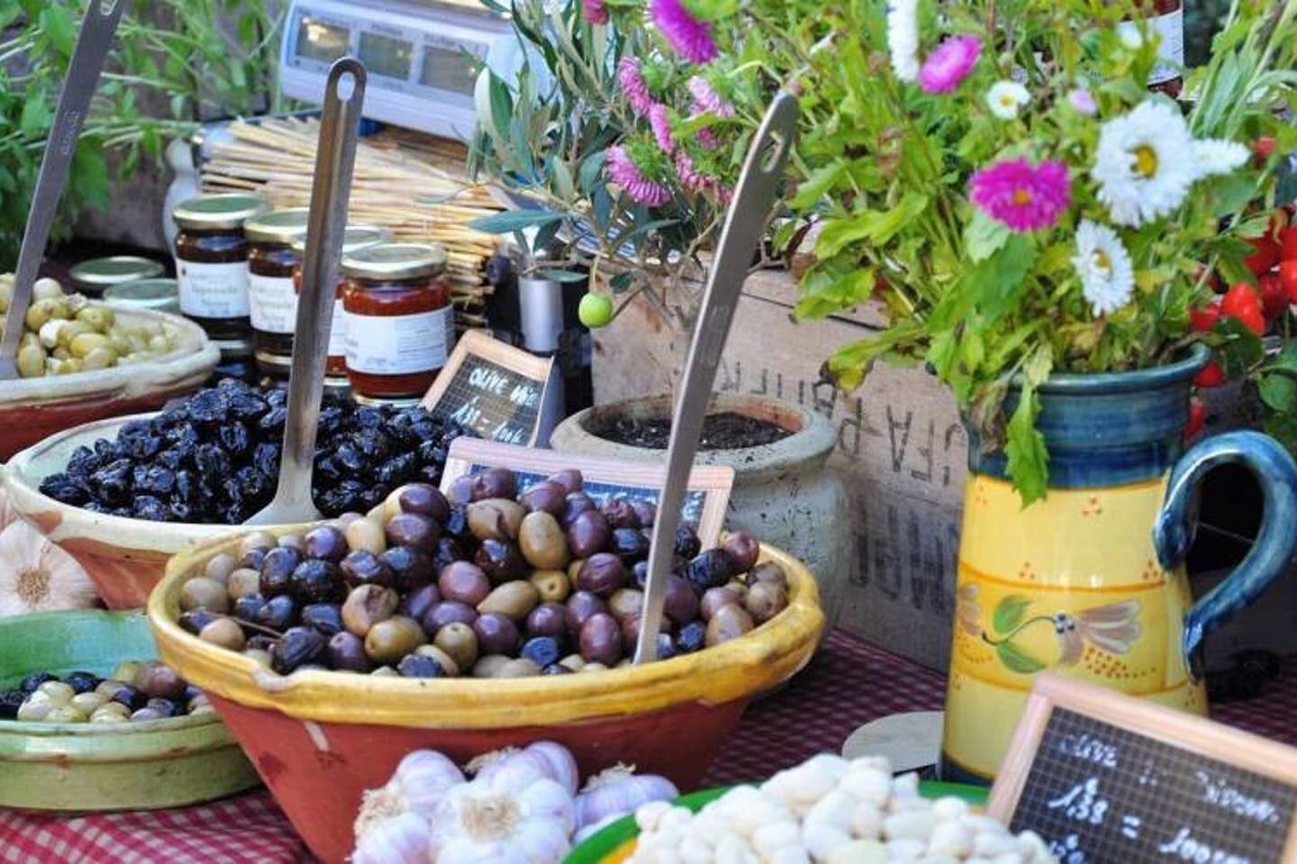 A market stall with various olives in bowls, a potted plant with flowers, and a chalkboard sign.
