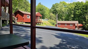 On aperçoit une cabane en bois rouge avec un porche, une petite construction en bois dotée d’un balcon, et une route au premier plan.