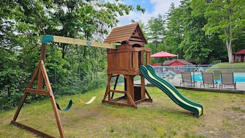 Une balançoire en bois avec un toboggan, entourée d’herbe et d’arbres.