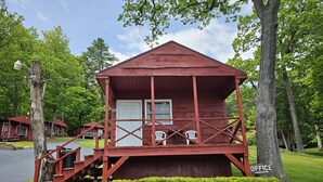 A small red wooden cabin with a porch and two white chairs. - The Gold Eagle Cottage 16 (Lake George)
