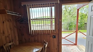 A wooden interior with a round table and chairs, a window with bars, and a view of a road and greenery outside. - The Gold Eagle Cottage 16 (Lake George)