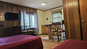 Una habitación con dos camas, un televisor montado en la pared, una kitchenette y una ventana con cortinas.