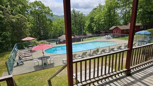 Una terraza de madera con zona de piscina, que incluye sillas de descanso y sombrillas, rodeada de vegetación y montañas al fondo.